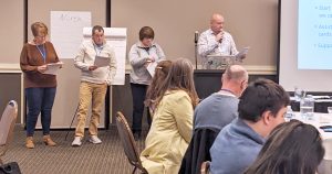 image of four people presenting in front of room. Man is holding a microphone. People in foreground are sitting at tables.
