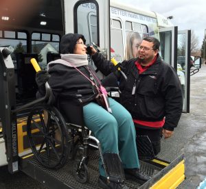 woman on a wheelchair lift entering a transit vehicle with the assistance of a male driver 